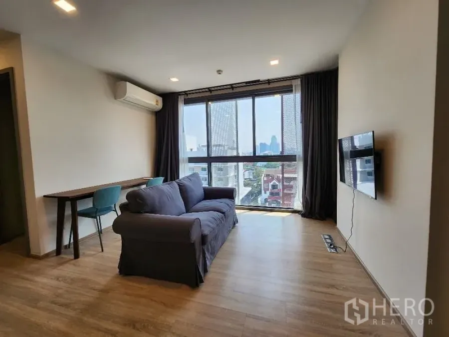 Condominium for rent in Khlong Toei, Bangkok - Bright living room with sofa, wall‑mounted TV, work desk and large window with city view.