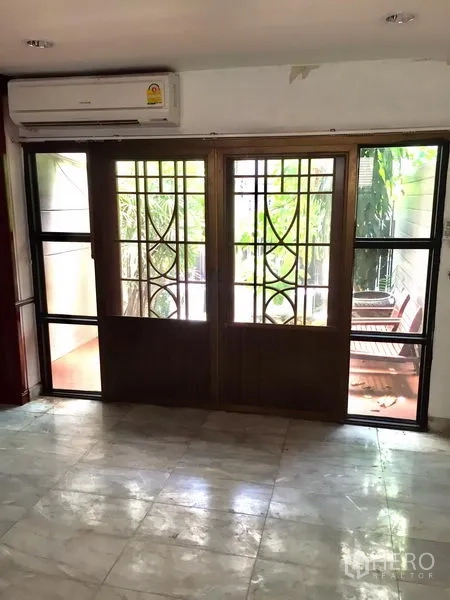 Townhouse for rent in Watthana, Bangkok - Ground-floor space with marble flooring and decorative metal-framed sliding doors to a terrace with greenery.