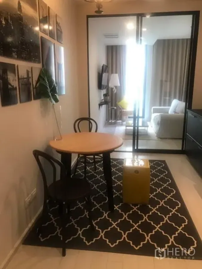 Condominium for rent in Pathum Wan, Bangkok - Compact dining nook with a round wooden table on a patterned rug, glass partition opening to a bright living area in a Pathum Wan condo near BTS Phloen Chit.