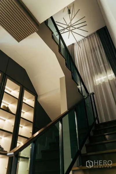 Detached House for sale in Sai Mai, Bangkok - Modern staircase with glass balustrade, tall curtains and a lit display cabinet beneath a chandelier.
