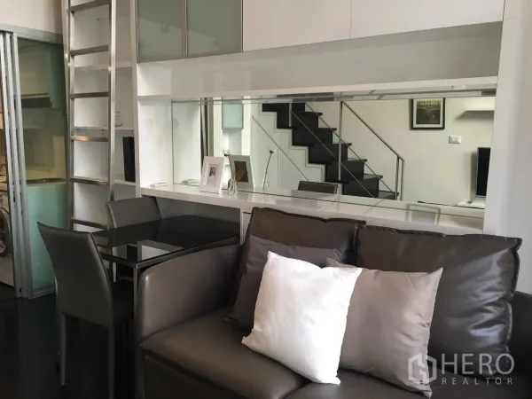 Condominium for rent in Khlong Toei, Bangkok - Modern living area with leather sofa, glass dining table, mirrored storage wall and sliding door to kitchen.