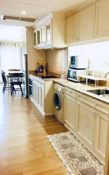 Condominium for rent in Khlong Toei, Bangkok - Galley kitchen with white cabinetry, countertop, sink, washing machine and appliances leading toward the dining area.