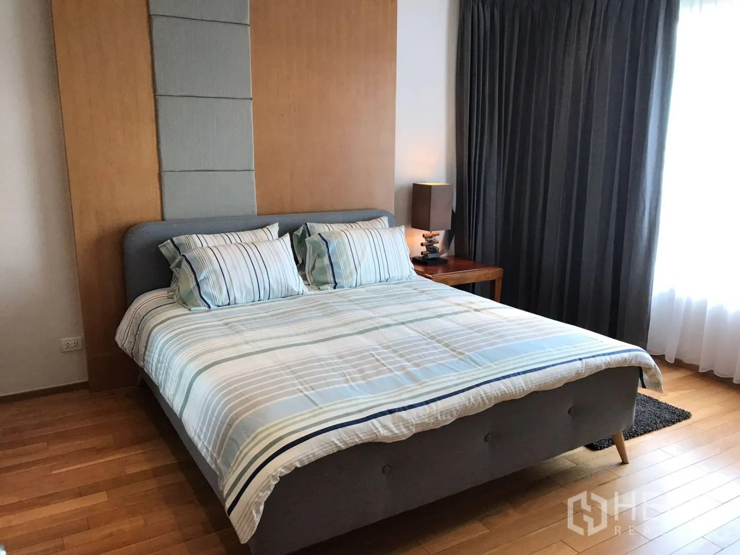Condominium for rent or sale in Khlong Toei, Bangkok - Bright bedroom with a king bed in striped linens, padded headboard, wooden flooring and blackout curtains.