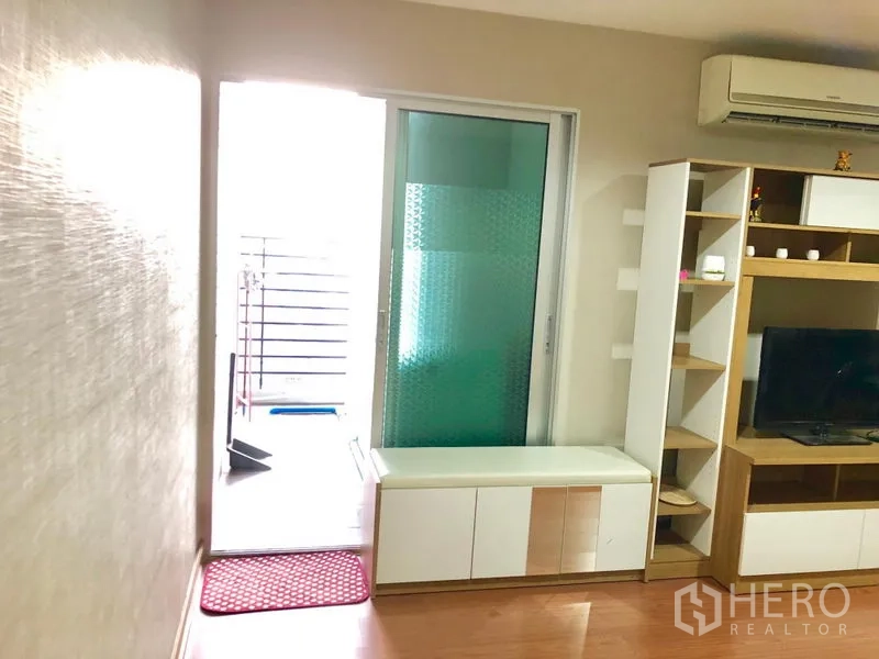 Condominium for rent in Huai Khwang, Bangkok - Living area with TV cabinet, open shelving, air conditioner, and sliding glass door to the balcony.