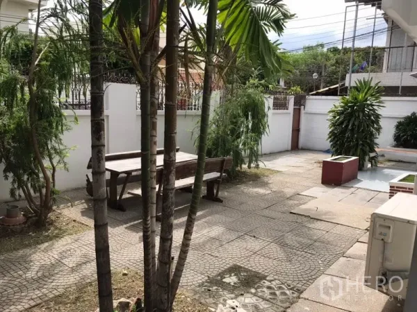 独立式住宅 出租在Phra Khanong, Bangkok - 前院铺石露台配木质长椅，棕榈与白色围墙环绕。