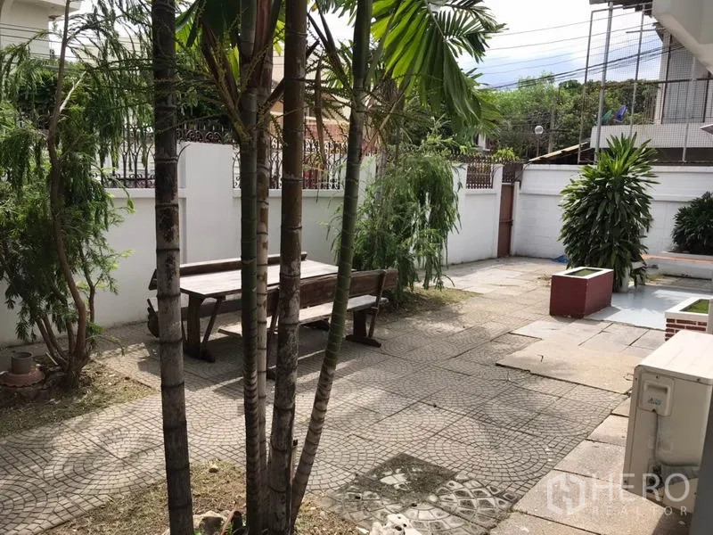 独立式住宅 出租在Phra Khanong, Bangkok - 前院铺石露台配木质长椅，棕榈与白色围墙环绕。
