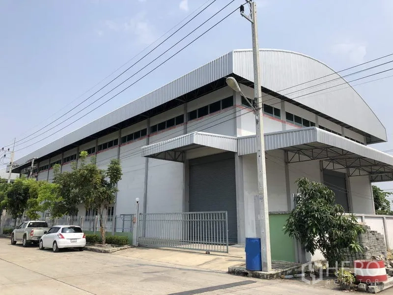 Factory for rent in Lam Luk Ka, Pathum Thani - Street view of a modern grey factory with curved metal roof, gated entrance and large roller shutter in Lam Luk Ka.