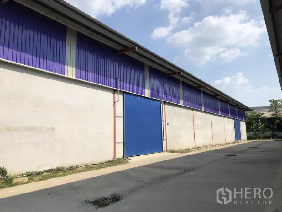 Warehouse for rent in Sai Noi, Nonthaburi - Side exterior of a long warehouse with purple metal cladding, a blue sliding steel door and a concrete driveway under a clear sky.