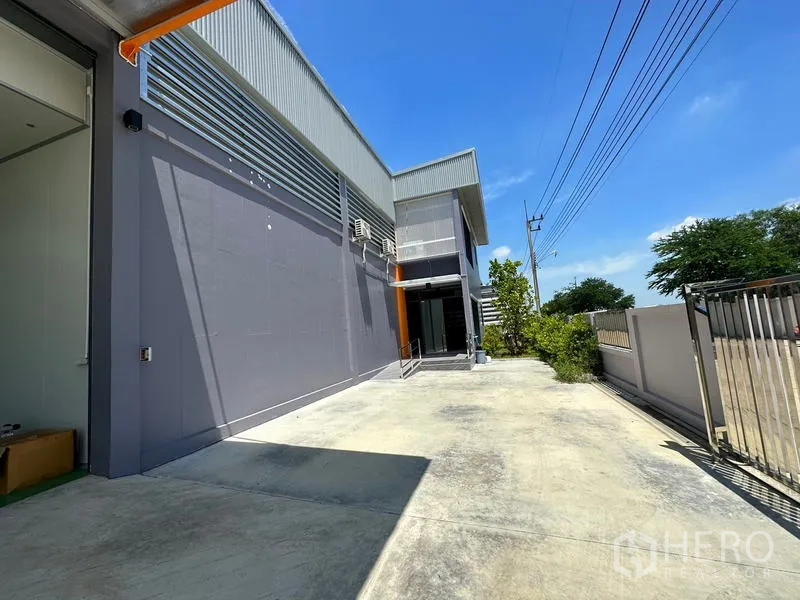 Factory for rent or sale in Lam Luk Ka, Pathum Thani - Gated side yard with long concrete driveway running along the factory wall.