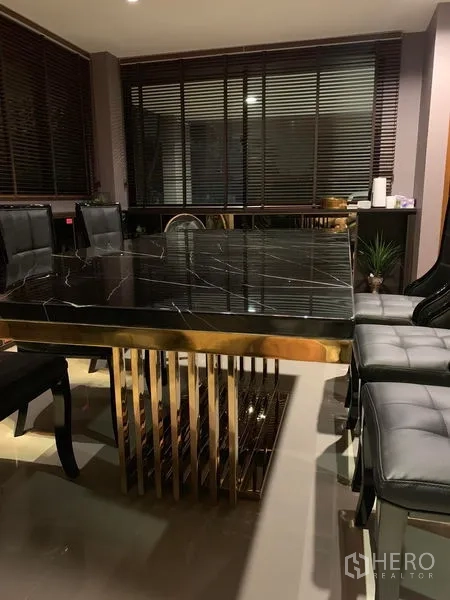 Townhouse for rent in Suan Luang, Bangkok - Elegant dining area with a black marble table on a gold base and leather chairs beside window blinds.