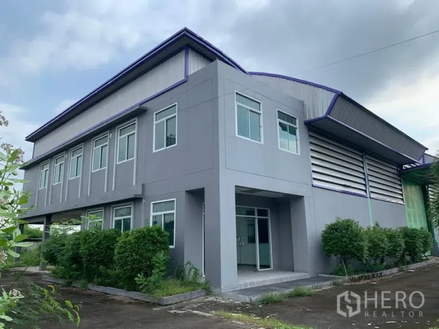 Warehouse for rent in Lam Luk Ka, Pathum Thani - Modern two-storey office attached to a grey warehouse with landscaped shrubs and driveway in Lam Luk Ka.