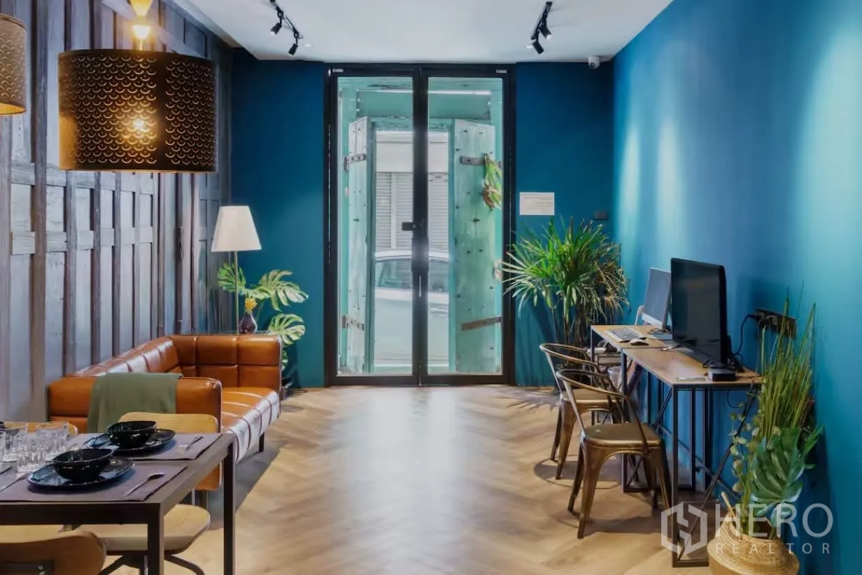 Townhouse for rent in Watthana, Bangkok - Open-plan living and workspace with leather sofa, dining set, indoor plants, and glass entry doors.