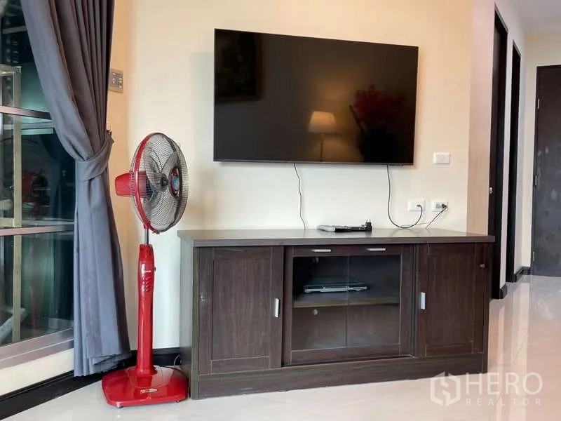 Apartment for rent in Khlong Toei, Bangkok - Wall-mounted TV above dark wood media cabinet next to full-length curtains.