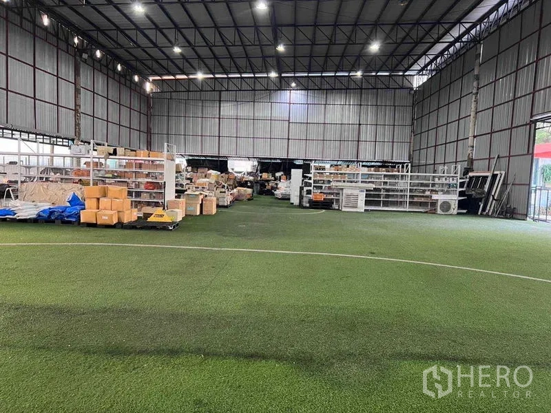Warehouse for rent in Lat Phrao, Bangkok - Wide open-span warehouse space with turf floor, metal roof, shelving and packed goods.