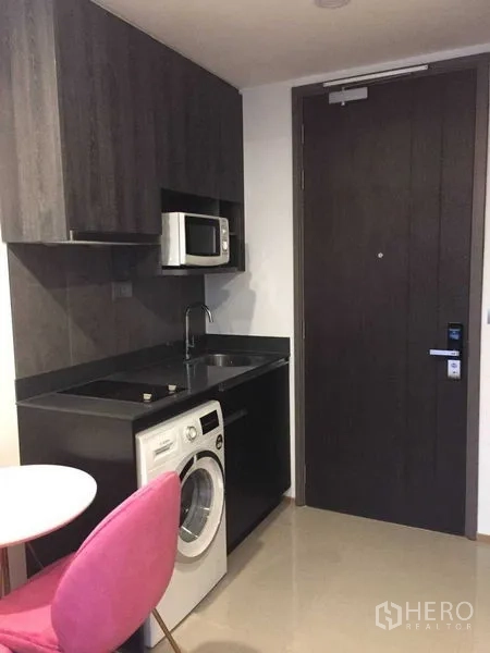 Condominium for rent in Bang Rak, Bangkok - Compact dark-tone kitchenette with sink, microwave and washing machine by the entry door.