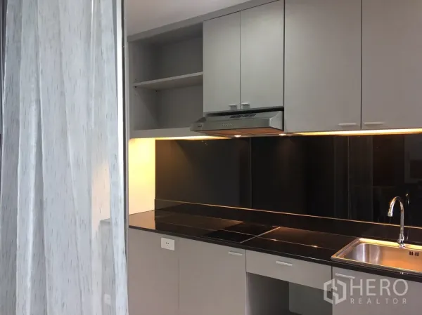 Condominium for sale in Phra Khanong, Bangkok - Modern kitchenette with grey cabinetry, black backsplash, induction hob, range hood and stainless-steel sink.
