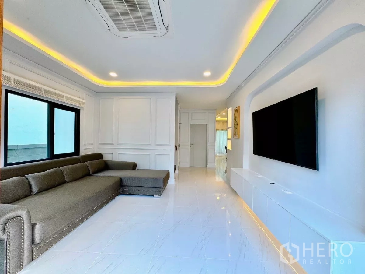 Detached House for rent in Bang Phli, Samut Prakan - Bright living room with L-shaped sofa, cove lighting, white tiled floors and a large TV wall.