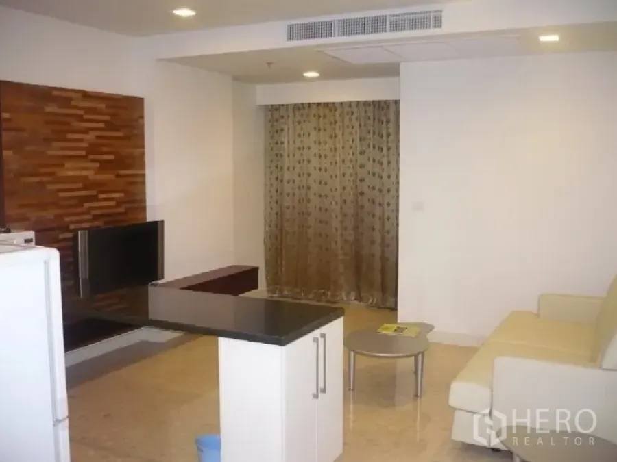 Condominium for rent in Khlong Toei, Bangkok - Bright Ekkamai condo living area with ivory sofa, round coffee table, TV wall with wood accents, island counter and curtains.