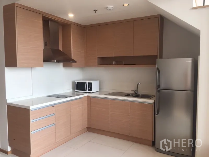 Condominium for rent in Khlong Toei, Bangkok - Modern L-shaped kitchen with light wood cabinets, range hood, cooktop, sink, microwave and refrigerator.