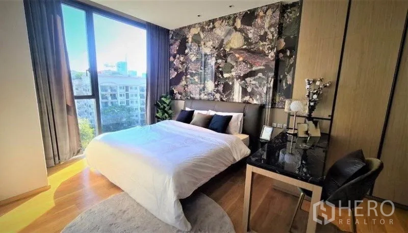 Condominium for rent in Khlong Toei, Bangkok - Sunlit bedroom with patterned headboard wall, large windows, work desk and greenery outside.