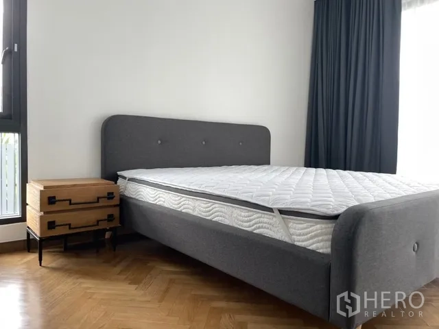 Detached House for rent in Watthana, Bangkok - Bright bedroom with a gray upholstered bed, wooden nightstand, and parquet flooring.