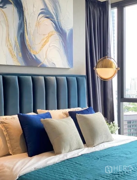 Condominium for rent - Close‑up of blue headboard with layered pillows beneath abstract artwork by a tall window.