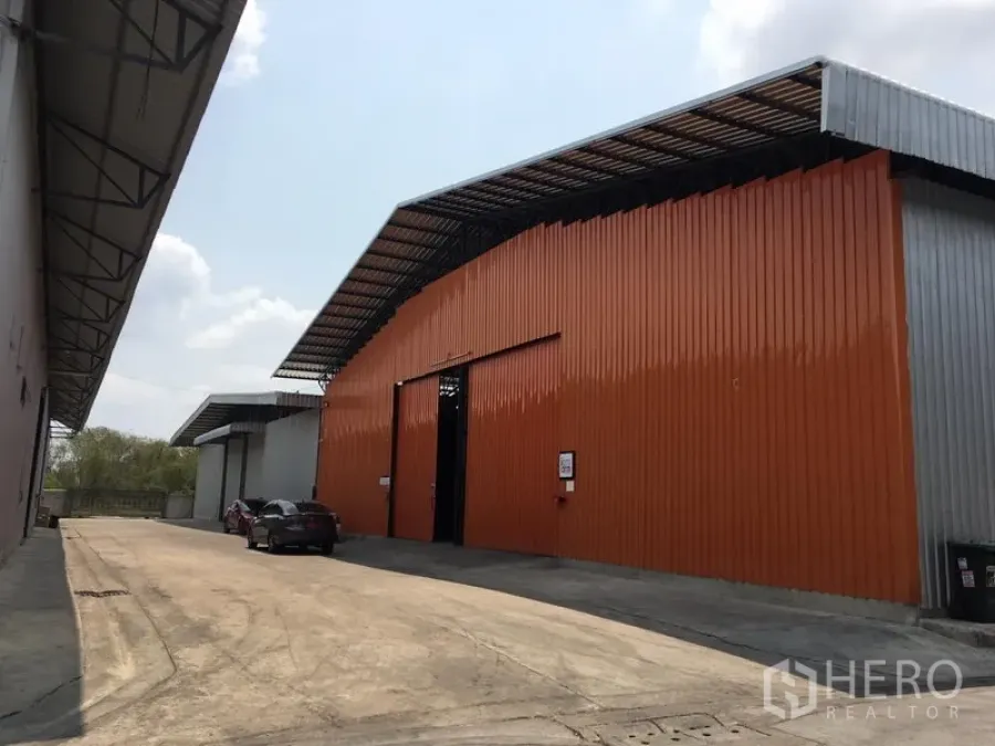 Warehouse for rent in Bang Kapi, Bangkok - Exterior view of a large orange and gray corrugated metal warehouse with a wide driveway in Bang Kapi.
