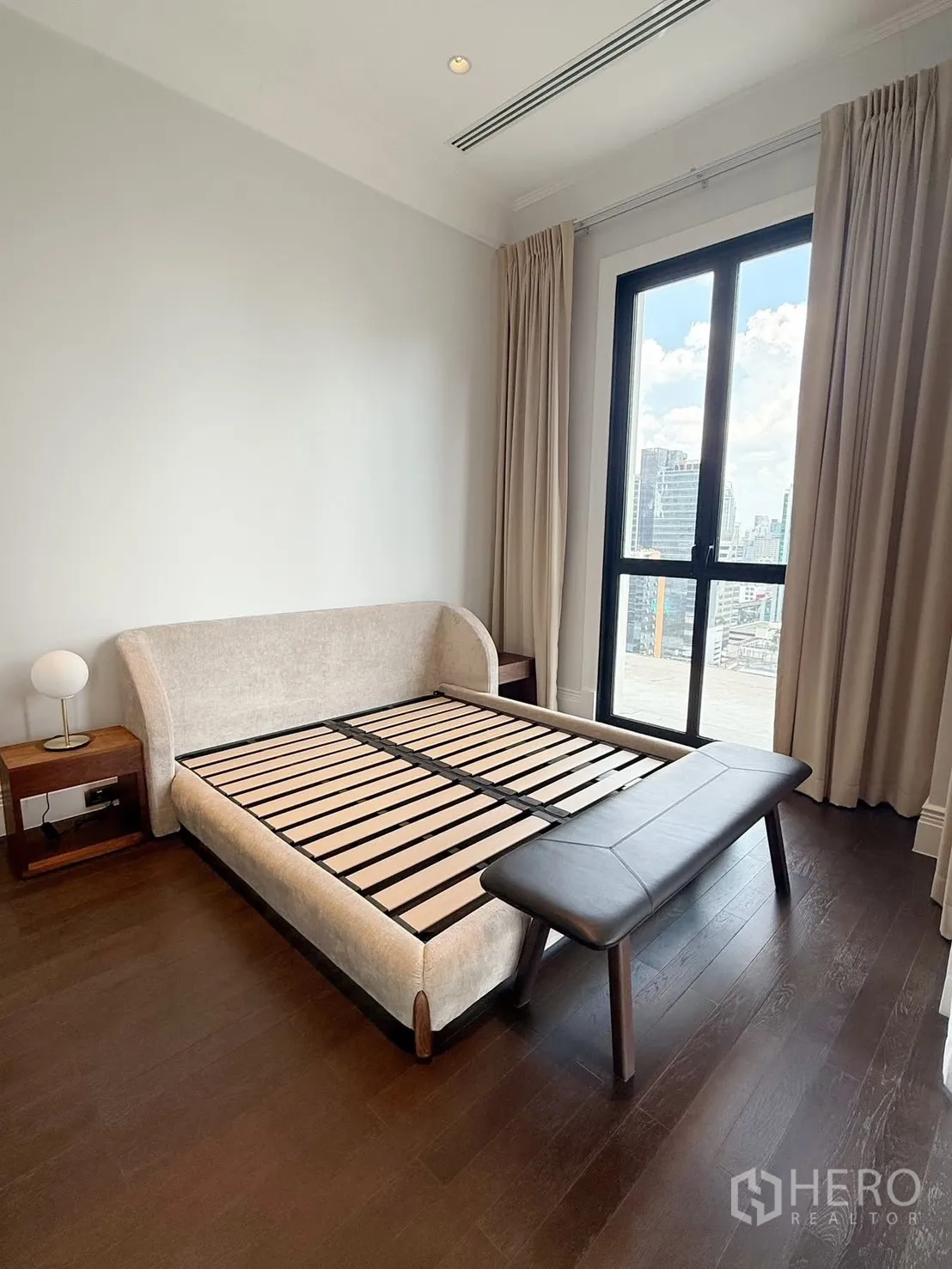Condominium for rent or sale in Pathum Wan, Bangkok - Bright bedroom with upholstered bedframe, bench, dark wood floors, and a large window facing the city.