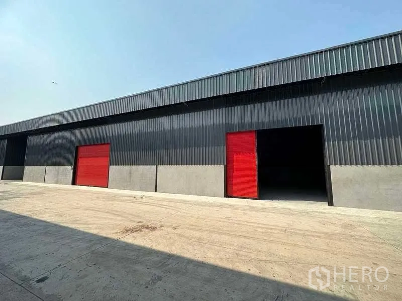 Warehouse for rent in Khlong Sam Wa, Bangkok - Row of black steel warehouses with two red roller shutters and a spacious driveway under clear sky.