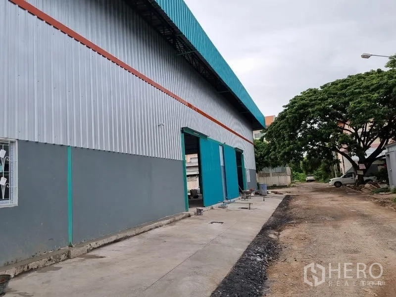 Warehouse for rent in Saphan Sung, Bangkok - Side view of the warehouse showing multiple bay doors and a concrete driveway along a tree-lined lane.
