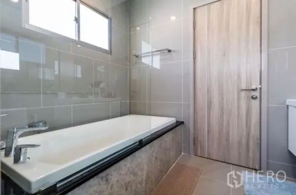 Detached House for rent - Modern bathroom with soaking bathtub, gray tiled walls, a window for light, and a wooden door.
