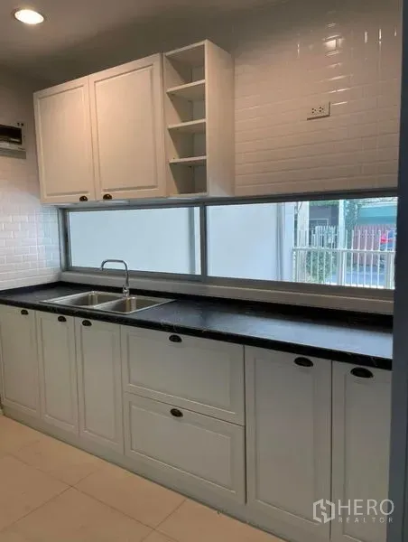 Townhouse for rent in Suan Luang, Bangkok - Bright galley kitchen with white shaker cabinets, subway tile backsplash, long black countertop and double sink beneath a wide window.