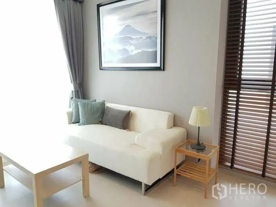 Condominium for rent or sale in Khlong Toei, Bangkok - Cozy living corner with white sofa, side table lamp and tall window blinds.