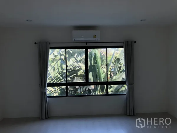Townhouse for rent in Bang Kapi, Bangkok - Bright bedroom with large window, grey curtains and air conditioner overlooking tropical greenery.