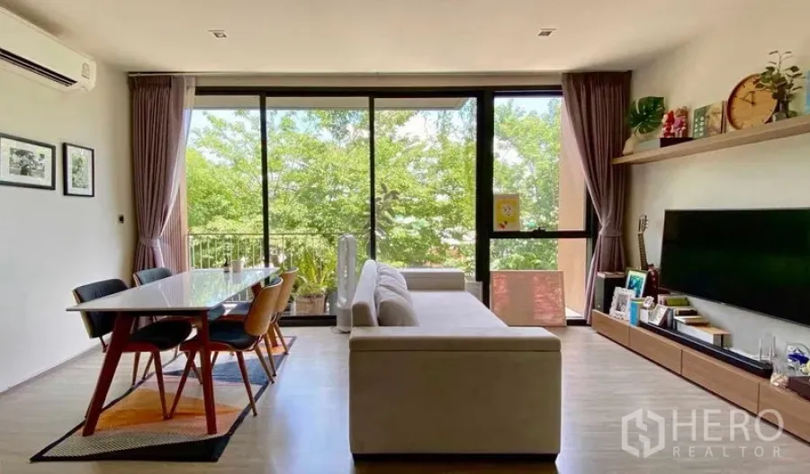 Condominium for sale in Watthana, Bangkok - Open-plan living and dining area with sofa and table facing full-height windows and a balcony with treetop views.