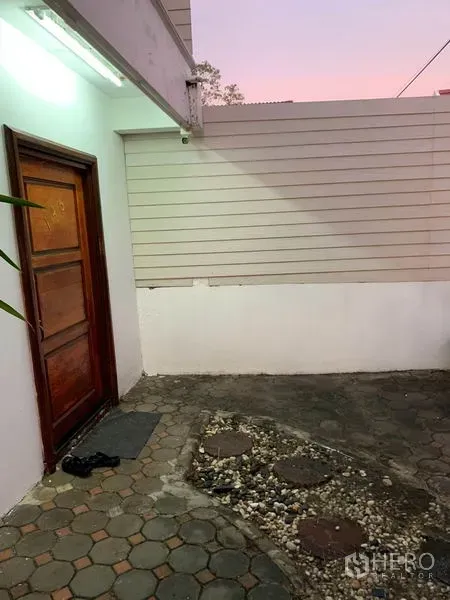 Townhouse for rent or sale in Phra Khanong, Bangkok - Front courtyard with pavers, white privacy wall and a wooden entrance door at dusk.