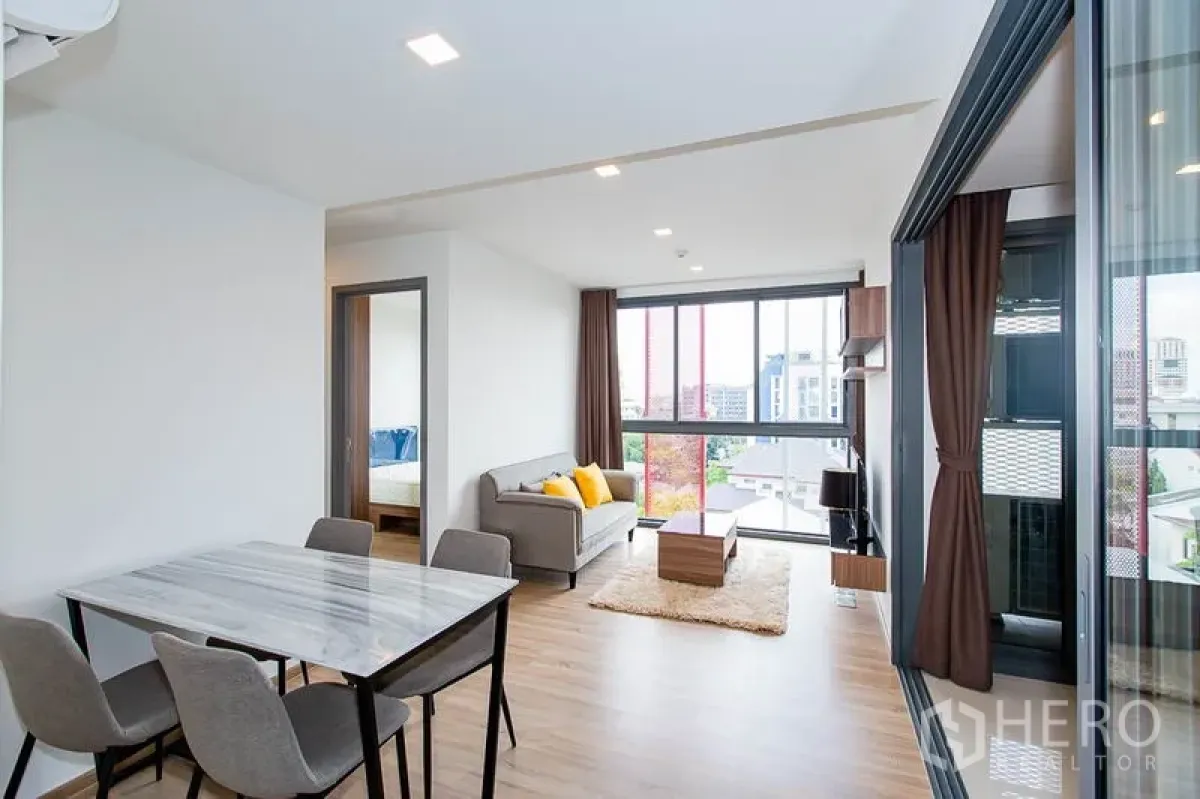 Condominium for rent in Watthana, Bangkok - Bright open-plan living and dining area with marble-top table, wood floors, sofa and full-height windows in a Watthana condo near BTS Ekkamai.
