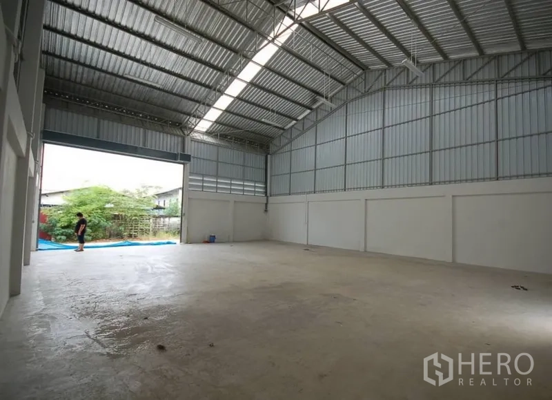 货栈 出租在Nonthaburi - 高挑金属屋顶的仓库内部，天窗采光，宽大卷闸门通向外部。