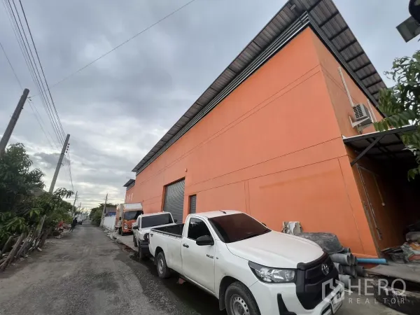 Warehouse for rent or sale in Lat Krabang, Bangkok - Side view of the orange warehouse along a narrow lane with parked pickup trucks and loading access.
