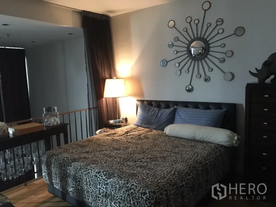 Condominium for rent or sale in Khlong Toei, Bangkok - Cozy master bedroom with tufted headboard, sunburst wall mirror, bedside lamp and dark curtains.