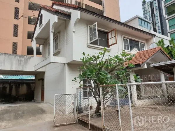 Detached House for rent or sale in Watthana, Bangkok - Two-storey detached house with tiled roof, gated driveway, covered parking and a mature tree.
