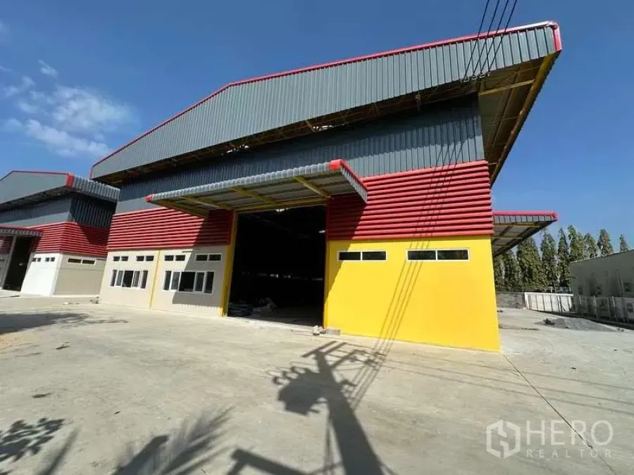Warehouse for rent in Lam Luk Ka, Pathum Thani - Colorful industrial warehouse exterior with large roller door, canopy, and wide concrete yard under blue sky in Lam Luk Ka.
