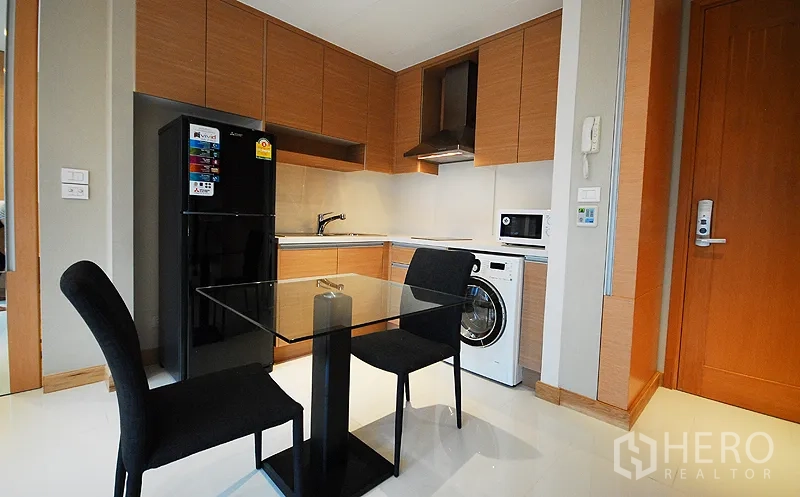 Condominium for rent in Khlong Toei, Bangkok - Modern kitchenette with wood cabinets, glass dining table, black refrigerator, range hood, microwave and in-unit washing machine.