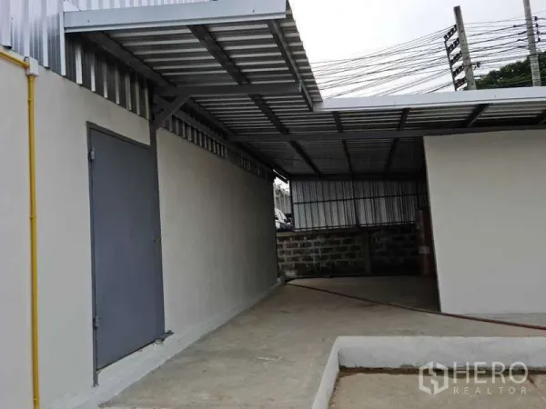 Warehouse for rent in Bang Bua Thong, Nonthaburi - Covered side service area with steel roof, solid door and utility lines beside the warehouse.