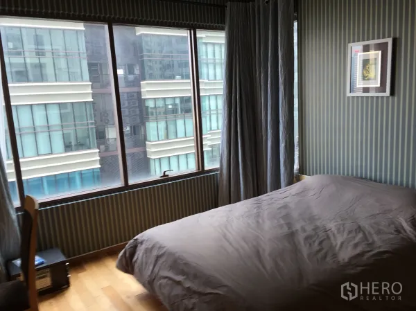 Condominium for rent or sale in Khlong Toei, Bangkok - Bedroom with large windows, striped wallpaper and city view from a mid-rise neighboring building.