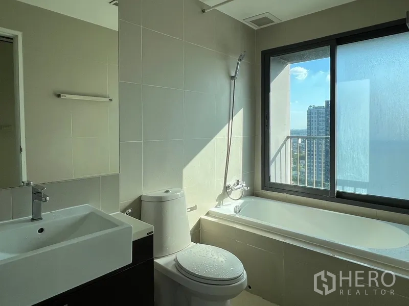 Condominium for rent in Khlong Toei, Bangkok - Modern bathroom with bathtub, vanity and toilet, plus a window offering city views.