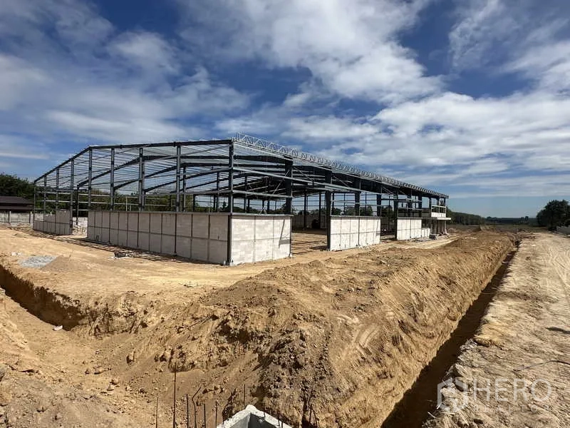 Warehouse for rent or sale in Nikhom Phatthana, Rayong - Closer exterior of the warehouse steel structure and concrete base walls beside a dirt site road.