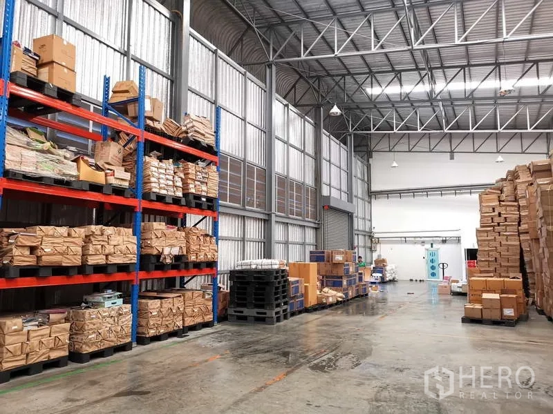 Warehouse for rent or sale in Nonthaburi - Warehouse interior with skylights, roller door, and stacked pallets beside steel racks.