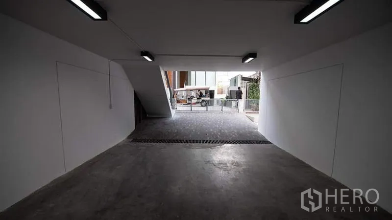 Townhouse for rent or sale in Phra Khanong, Bangkok - Covered parking bay beneath the townhouse with lighting and a driveway to the street.