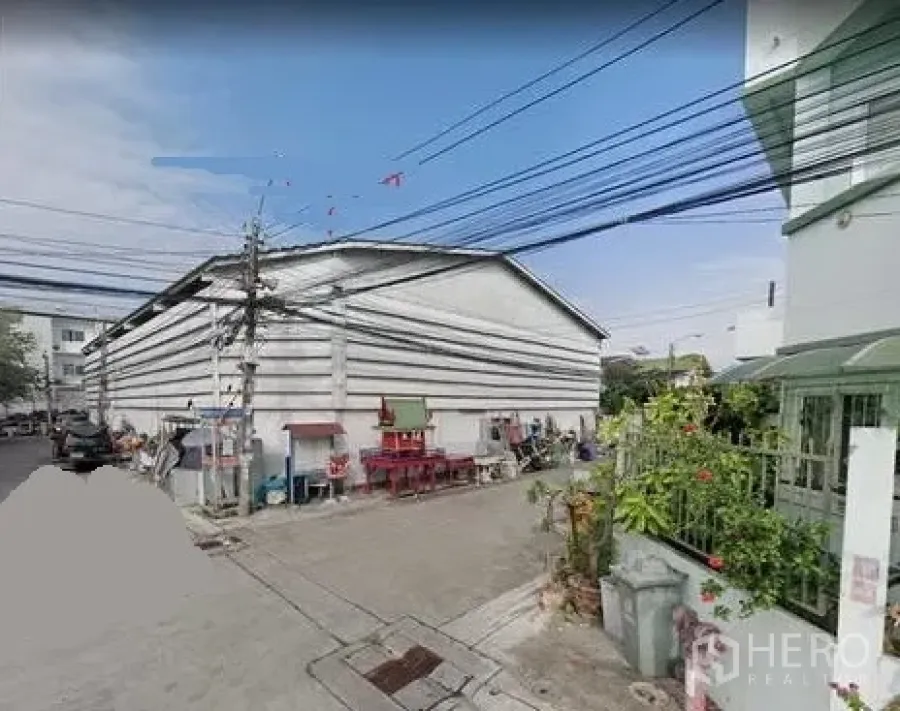 Warehouse for rent in Prawet, Bangkok - Street view of a large gable-roof warehouse with white siding on a quiet lane in Prawet, Bangkok, with power lines and nearby houses.